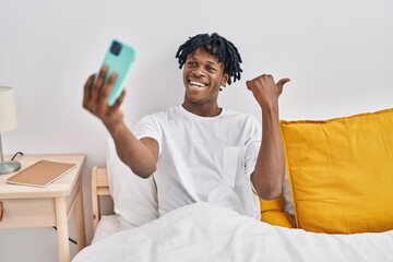 Young african man with dreadlocks taking a selfie on the bed pointing thumb up to the side smiling happy with open mouth