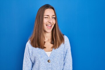 Young woman standing over blue background winking looking at the camera with sexy expression, cheerful and happy face.