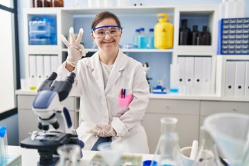 Hispanic girl with down syndrome working at scientist laboratory smiling positive doing ok sign with hand and fingers. successful expression.