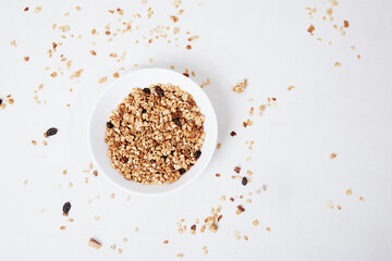 A bowl of granola sits on a white table with scattered granola on it.