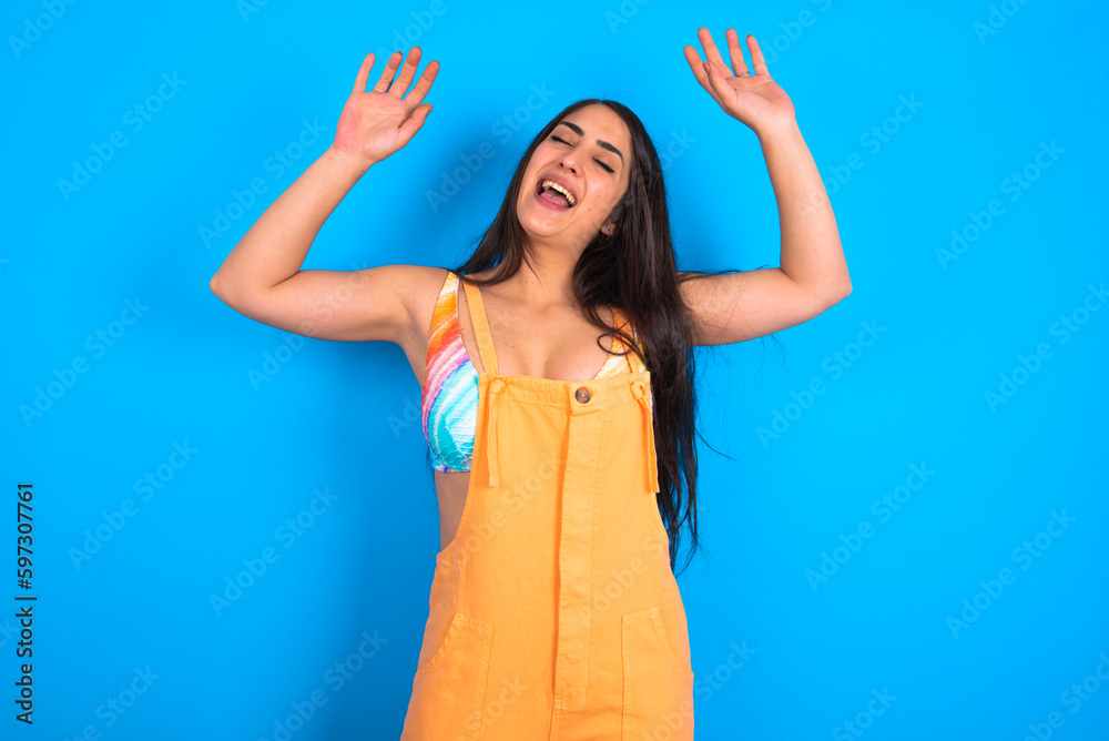 Wall mural Photo of upbeat beautiful brunette woman wearing overall  over blue background has fun and dances carefree wear being in perfect mood makes movements. Spends free time on disco party