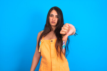 beautiful brunette woman wearing overall  over blue background looking unhappy and angry showing rejection and negative with thumbs down gesture. Bad expression.