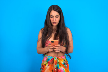 Focused beautiful brunette woman wearing bikini over blue background use smartphone reading social...
