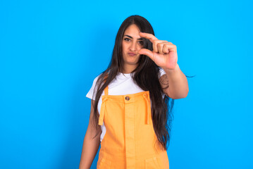 beautiful brunette woman wearing overall  over blue background purses lip and gestures with hand, shows something very little.