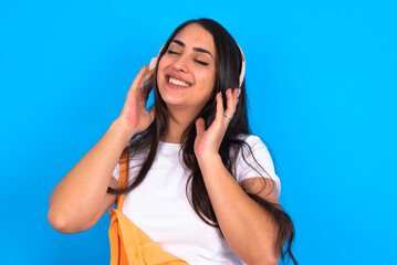 beautiful brunette woman wearing overall  over blue background with headphones on her head, listens to music, enjoying favourite song with closed eyes, holding hands on headset.