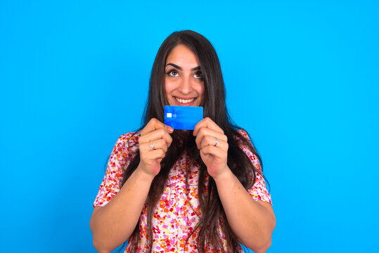 Photo of cheerful beautiful brunette woman wearing floral dress over blue background hold debit card look empty space