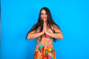 beautiful brunette woman wearing swimwear over blue background praying with hands together asking for forgiveness smiling confident.