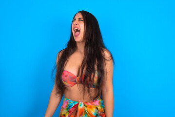 beautiful brunette woman wearing swimwear over blue background angry and mad screaming frustrated...