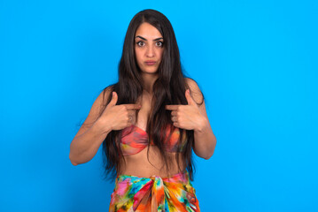 Embarrassed beautiful brunette woman wearing swimwear over blue wall indicates at herself with puzzled expression, being shocked to be chosen to participate in competition, hesitates about something