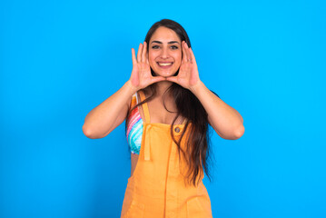 brunette woman wearing orange overalls over blue studio background Pleasant looking cheerful, Happy reaction