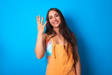 brunette woman wearing orange overalls over blue studio background smiling and looking friendly, showing number four or fourth with hand forward, counting down