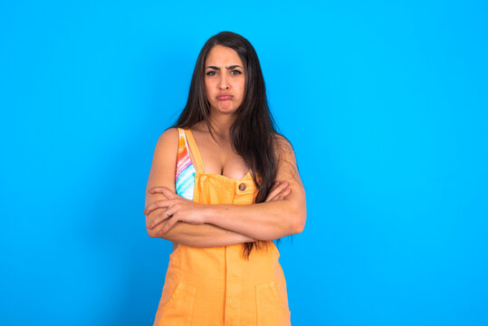 Gloomy dissatisfied brunette woman wearing orange overalls over blue studio background looks with miserable expression at camera from under forehead, makes unhappy grimace