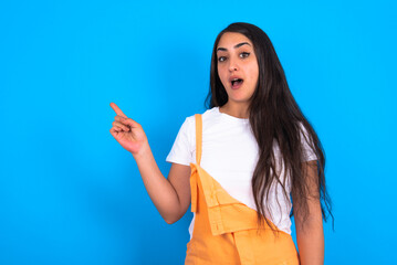 Emotive brunette woman wearing orange overalls over blue studio wall keeps jaw dropped from shock demonstrates amazing promo points right on blank space demonstrates big shopping sale. Advertisement