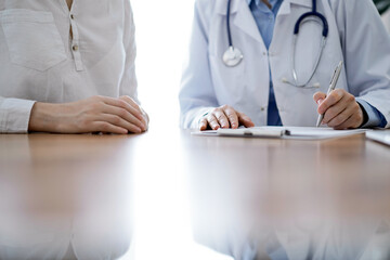 Doctor and patient sitting near each other at the wooden desk in clinic. Female physician's filling...