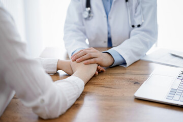 Doctor and patient sitting at the wooden table in clinic. Female physician's hands reassuring woman. Medicine concept