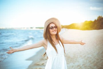 Fototapeta na wymiar Happy smiling woman in free happiness bliss on ocean beach standing with a hat, sunglasses, and open hands. Portrait of a multicultural female model in white summer dress enjoying nature