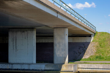 Cement concrete pillar structure of urban bridge crossing over small river, canal or ditch with green grass meadow, Underneath of classic style bridge under blue sky and white clouds, Netherlands.