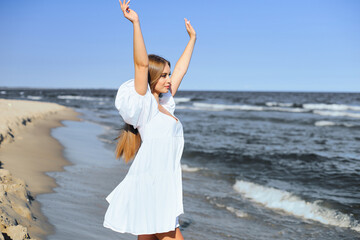 Happy, beautiful woman on the ocean beach standing in a white summer dress, raising hands