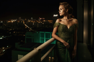 Attractive woman with a light tan complexion wearing a green evening gown and standing on a balcony overlooking a city at night, generative ai