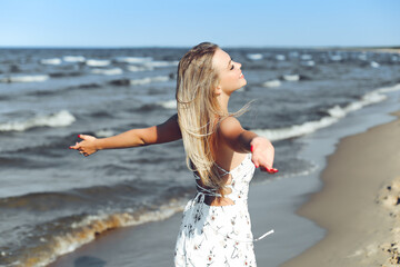 Fototapeta na wymiar Happy blonde beautiful woman on the ocean beach standing in a white summer dress, raising hands