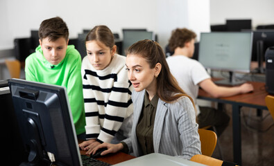 Fototapeta premium Friendly female teacher and positive teen students looking at monitor screen during lesson in computer lab at school library ..