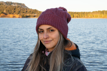 Portrait of a smiling young woman wearing a coat and purple hat with the lake and mountains in the background.