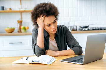 Frustrated african american curly young woman, student or freelancer, sit in a kitchen at a table with a laptop, disappointment look at screen, tired of studying or working online, having a headache