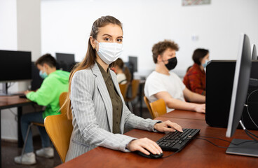 Positive female teacher wearing face mask working on computer in library. Concept of adult self-education during pandemic