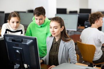 Interested teenagers studying with young female teacher in information technology class at college, sitting around one computer