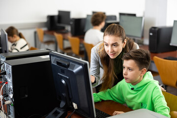 Positive female teacher helping focused preteen schoolboy studying in college library computer lab..