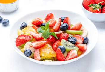 Fresh summer fruit and berry salad with strawberries, blueberries, banana, kiwi, orange, mint leaves and honey dressing, white table background, top view