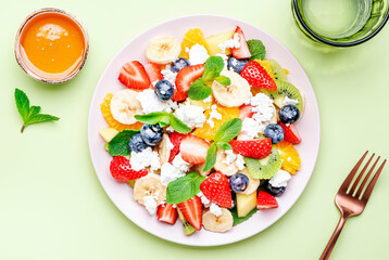 Summer fruit and berry salad with strawberries, blueberries, banana, soft cheese and mint leaves, green background, top view