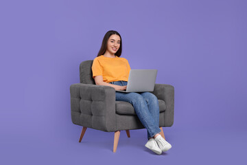 Smiling young woman with laptop sitting in armchair on lilac background, space for text