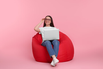 Smiling young woman with laptop sitting on beanbag chair against pink background