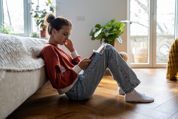 Teens and cyberbullying. Upset teen girl sitting on floor near bed using smartphone at home,...