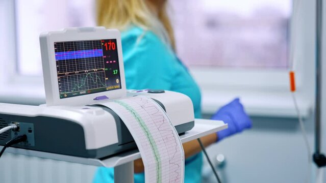 Cardiotocography device recording the fetal heart rate well-being. Female nurse wearing blue uniform standing at backdrop in blur.