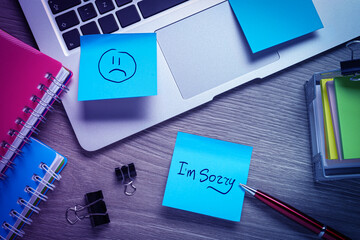 Sticky notes with word Sorry and drawn sad face on workplace. Laptop and stationery on wooden table, flat lay
