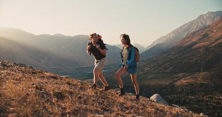 Caucasian couple hiking together with backpacks, helping each other on their way up the mountains - freedom, active lifestyle concept  - obrazy, fototapety, plakaty