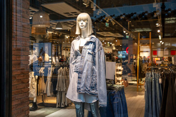 A female mannequin in a denim jacket in a glass showcase of a fashionable youth clothing store....