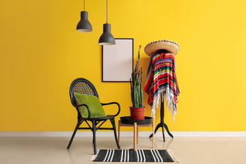 Rack with Mexican clothes, armchair and cactus on table near yellow wall