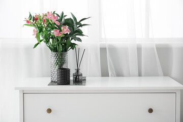 Reed diffuser and vase with beautiful flowers on chest of drawers near window