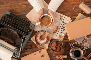 Woman with cup of coffee, typewriter, newspaper and pastry on wooden table