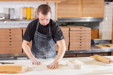 The chef prepares pizza. Raw pizza ready to bake. Cook in a blue apron in the kitchen. with a shovel in his hands. boxes for food delivery on background.