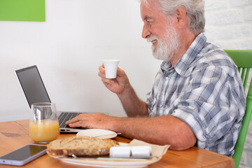 Bearded senior caucasian man sitting at the table in coffee shop using laptop holding an espresso coffee cup. Elderly man at breakfast time while typing on computer