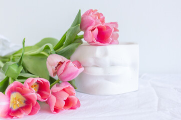 Pink tulips bouquet and plaster statue of lips lying on white wrinkled blanket background copy space