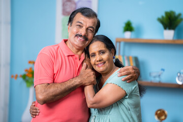 Fototapeta premium Happy Indian senior couple closely standing by embracing each other by looking camera at home - concept of family bonding, relationship and