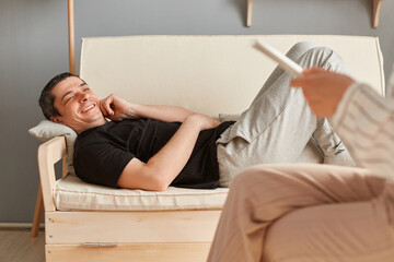 Cheerful satisfied young man in casual clothing laying on couch at counselor office, talking with...
