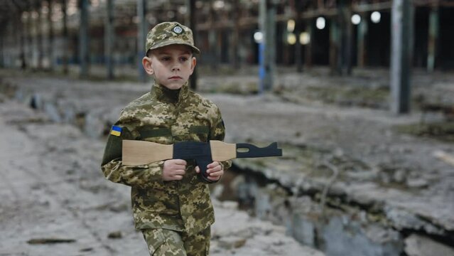 Little boy in military uniform going near destroyed plant holding a toy weapon against a background of devastation place. Soldier. Infantry. Storming the building. People and War Concept