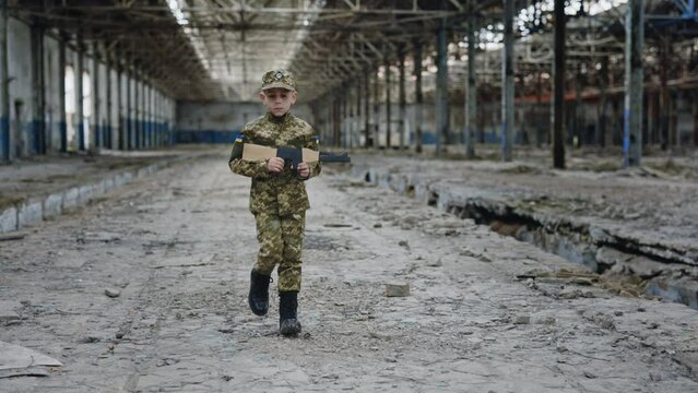 Ukrainian Little Soldier is Walking Holding a Toy Weapon Inside Destroyed Plant. Male Kid Wearing Military Wear. War. Patriot. Boy Playing War Games