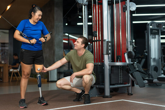 Asia Woman with prosthetic leg exercising on multi station for arm and caucasian trainer support at fitness or gym	